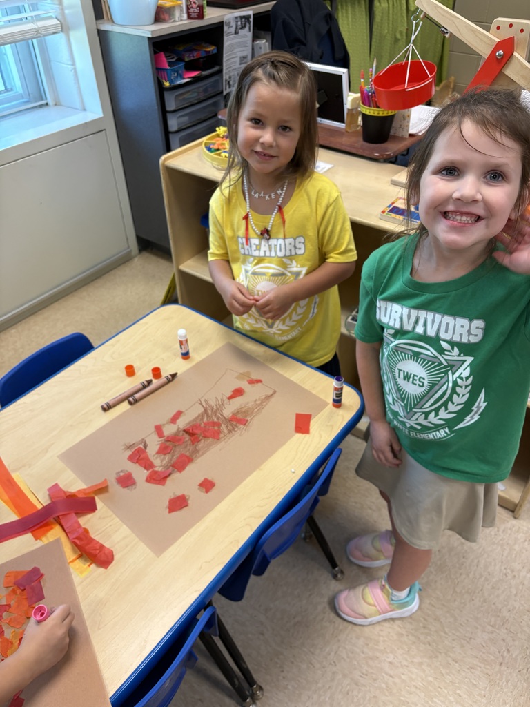 Two children standing by a desk with art supplies. A table displays a colorful art project.