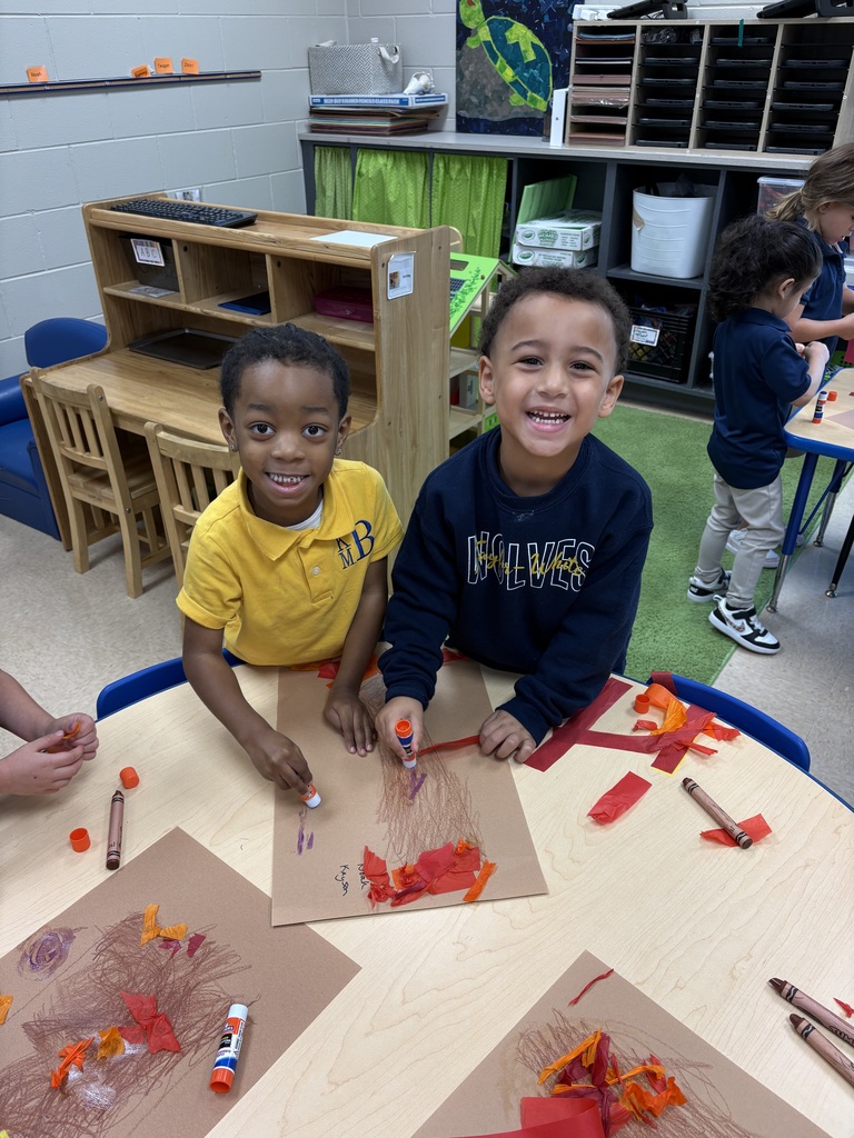 Two boys in a classroom, one in yellow, one in blue, sitting at a table creating art with orange paper.