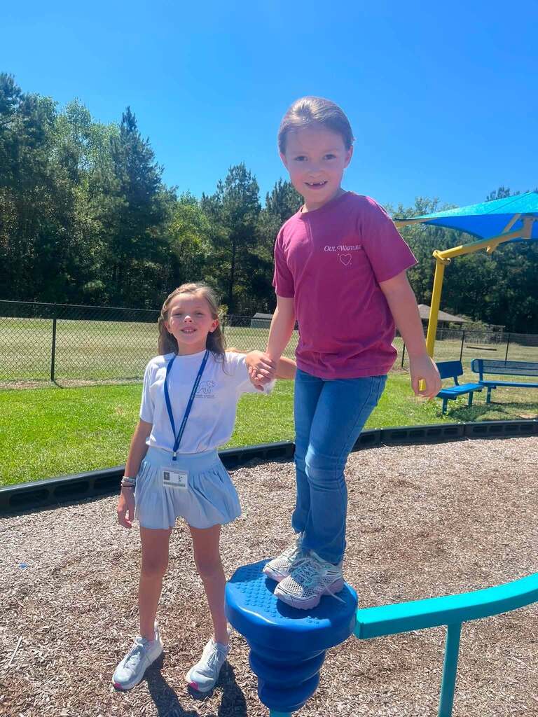 A fifth grader on the ground holds the hand of a kindergartener standing on a piece of playground equipment