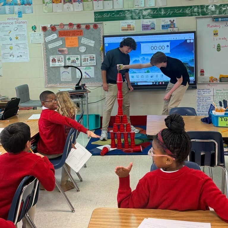 Two high school students stack plastic cups while a class of elementary students watch