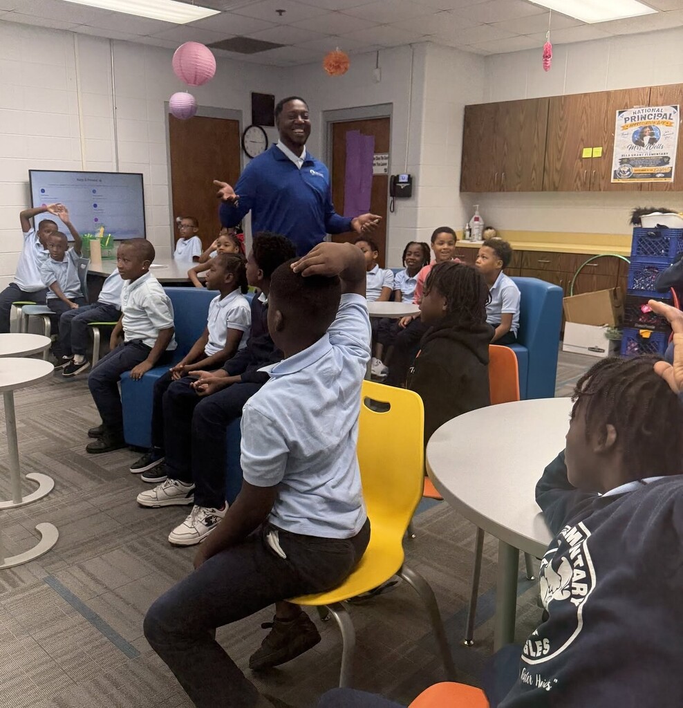 A man speaking in a classroom full of students, some of whom have their hands raised