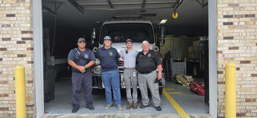 Four men stand in a fire station garage with a truck. Yellow poles are on the sides.