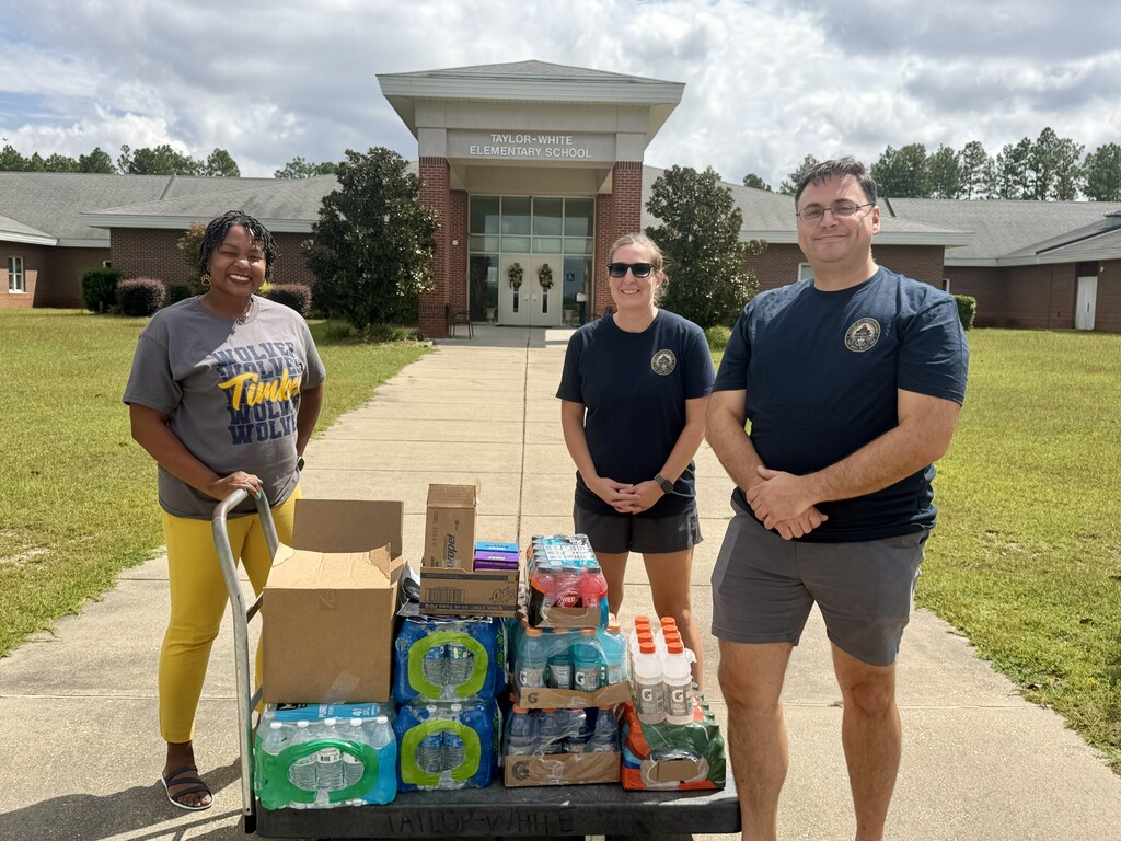 Three people pose near a cart full of bottled water in front of a building.