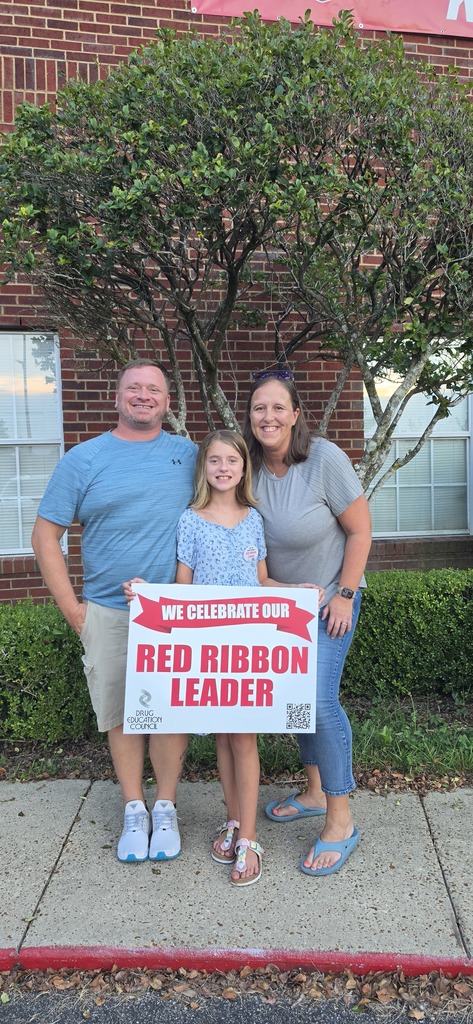 A family stands on a sidewalk with a sign reading "Red Ribbon Leader" in front of a brick building.