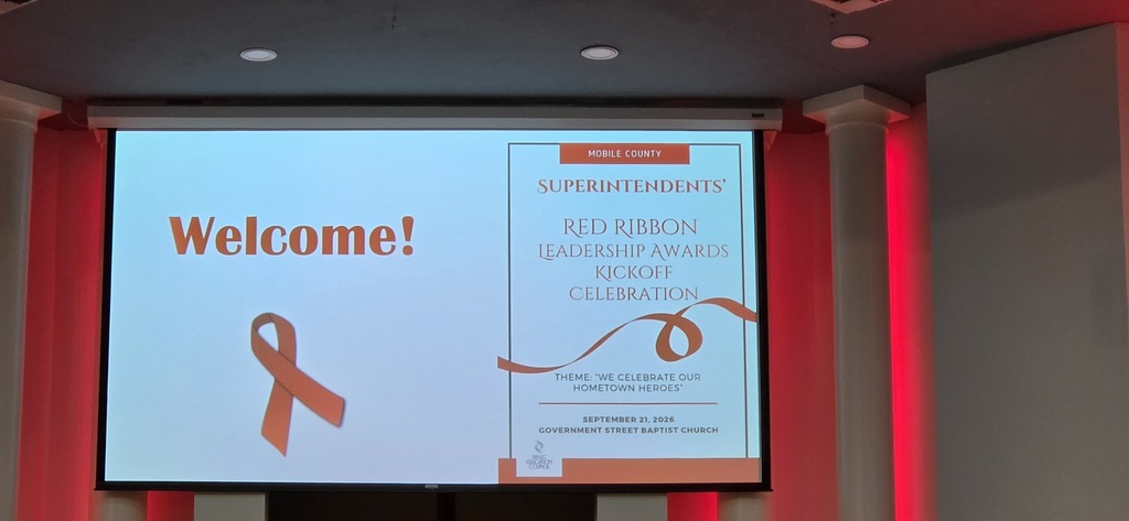 A projector screen displays "Welcome!" with a red ribbon logo, in a room with red lights and white walls.
