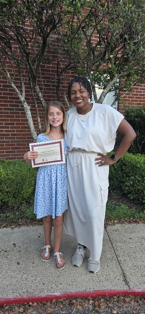 A young girl in a blue dress holds a framed award. Next to her, an adult woman in a white dress stands on a sidewalk.