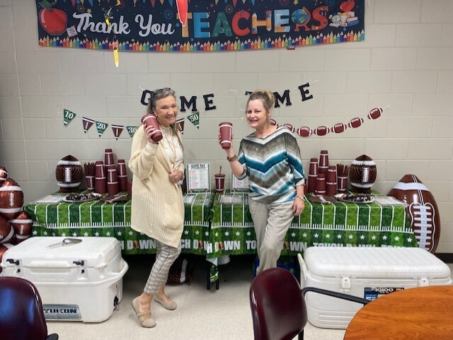 Two women stand in a room with a football-themed setup, featuring a "Thank You Teachers" banner and various footballs.