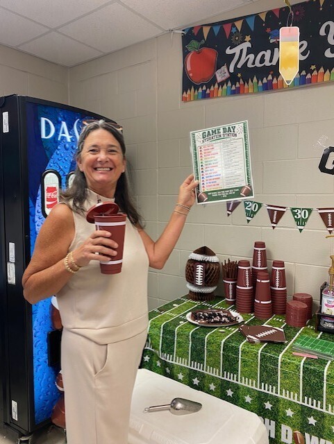 A smiling woman stands in front of a vending machine, holding a cup. Behind her, a table displays football-themed items.