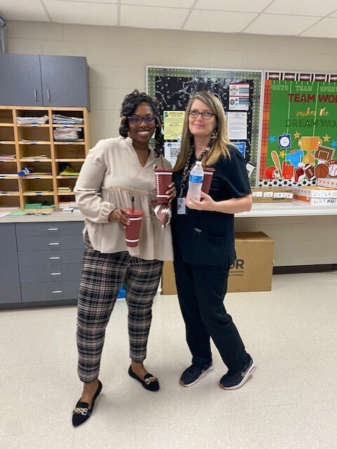 Two women in a room hold cups, one in each hand. One wears a white blouse, plaid pants, and black shoes. The other wears a black shirt, pants, and sneakers.