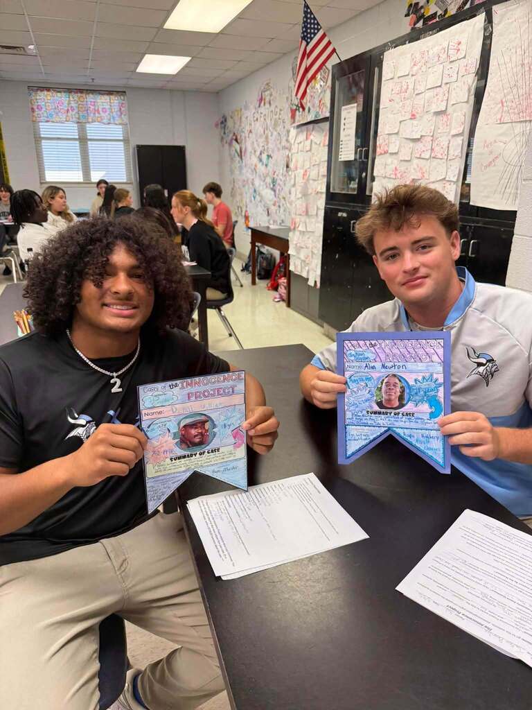 Two students sitting at a table in a classroom. One holds a certificate and the other a framed photo.