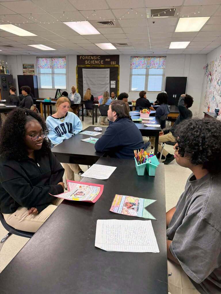 Several students sit around a long table, with papers and pencils in front of them. The room has windows and fluorescent lights.