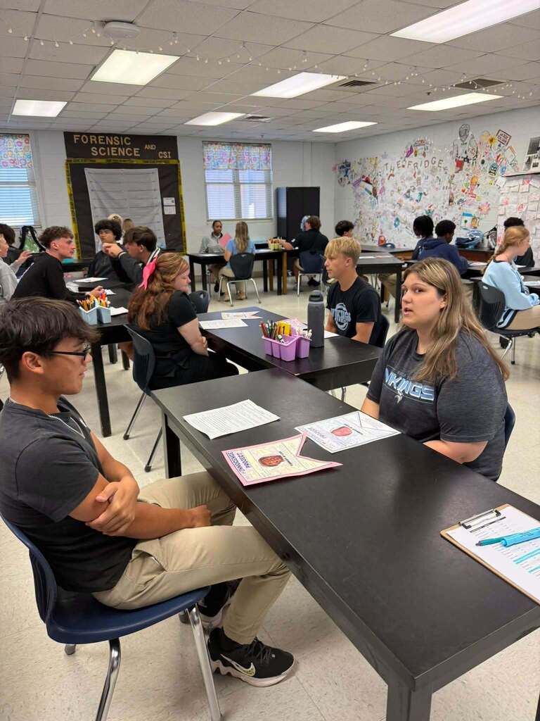 A classroom with students seated at desks. A teacher and student sit at a table, working on papers.