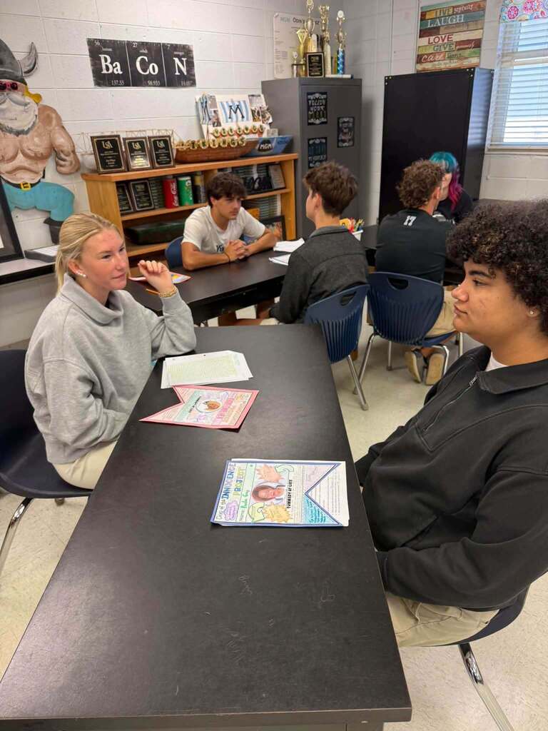 A classroom with students seated at desks; one student is conversing with another. Tables have books and papers.