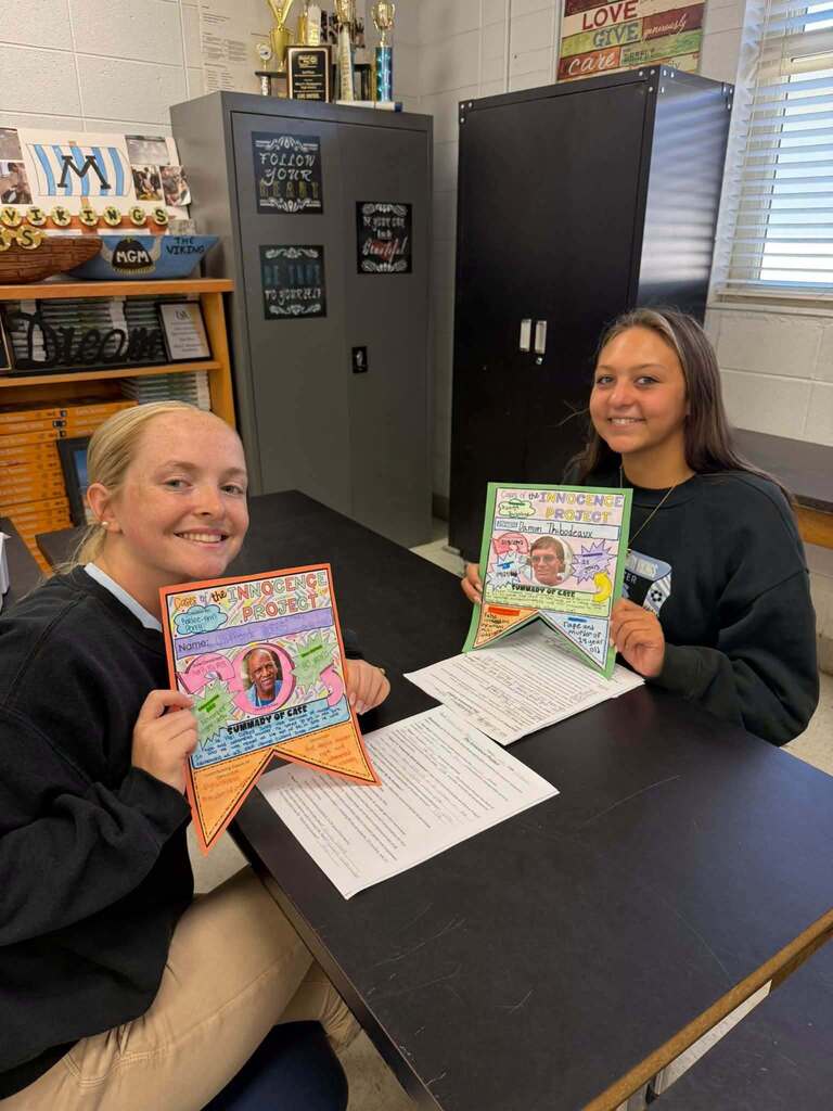 Two women seated at a table hold up homemade posters, smiling. Behind them is a classroom with storage cabinets and a bulletin board.