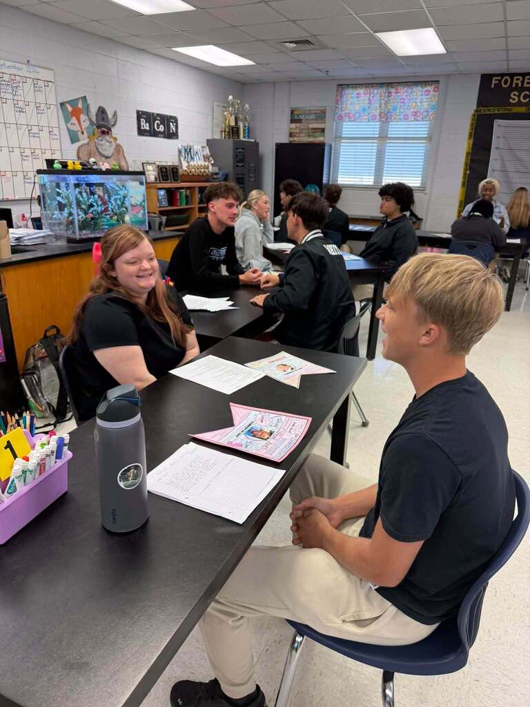 Students sit at a table with papers, one smiling, another with short hair, in a classroom with bright lighting.