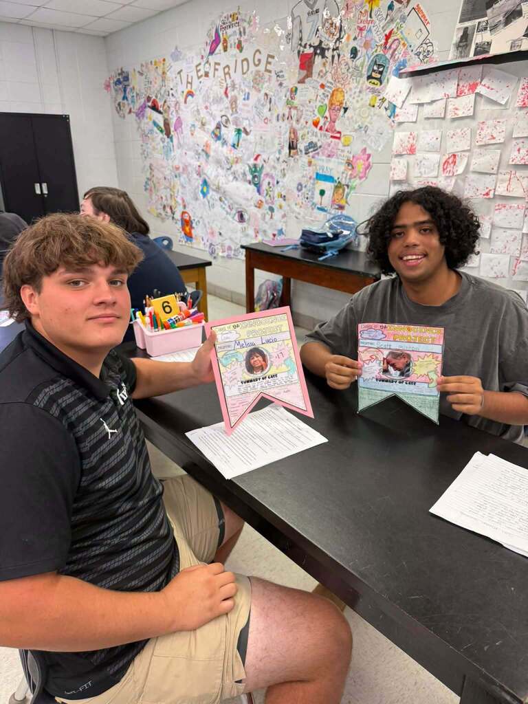 Two individuals seated at a desk hold up a certificate with a picture and banner. Behind them, a bulletin board has various notes.