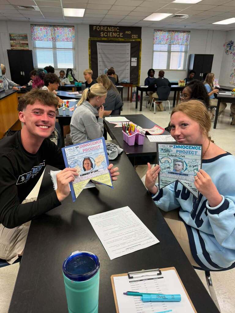 Two individuals in a classroom sit at a desk, holding up printed certificates. Other students are seated at tables in the background.