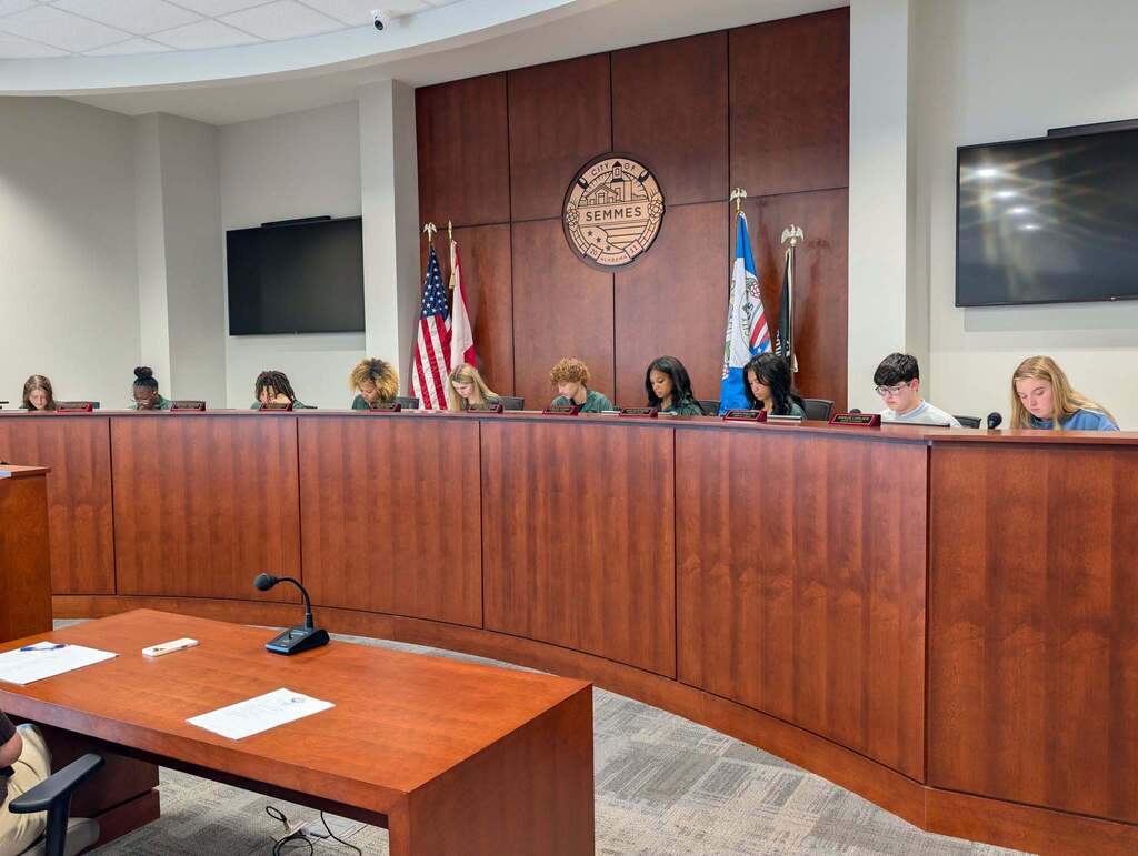 A council meeting with people seated around a curved wooden desk. Flags and monitors on the wall.