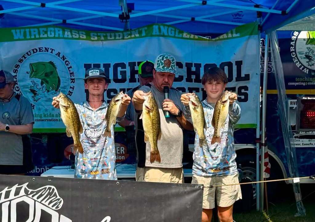 Three young men holding up fish, standing under a banner. A man stands beside them, holding a microphone.