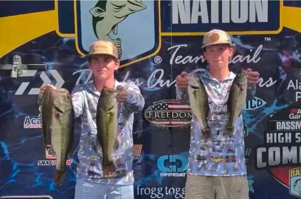 Two men in hats, holding fish, stand before a banner with logos, text, and a fish graphic.