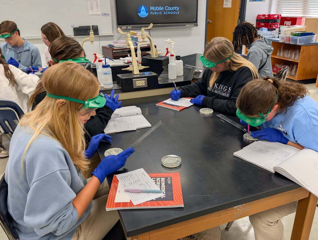 Students in goggles and gloves conduct a lab in a classroom, seated at a table with open books and science equipment.