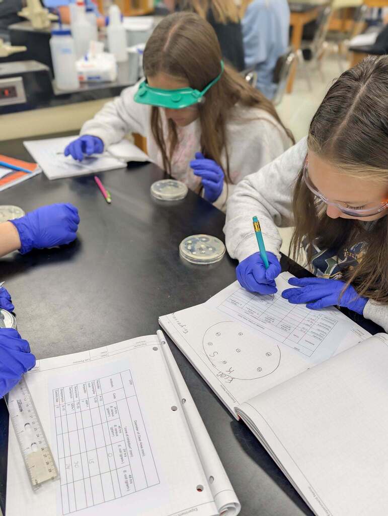 Three students in a science lab, wearing goggles and gloves, work at a table with lab equipment.
