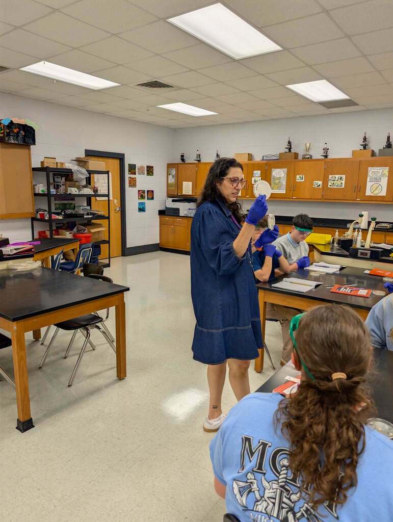 A teacher in a blue dress stands in a classroom, speaking to students seated at tables. The room has cabinets and overhead lighting.