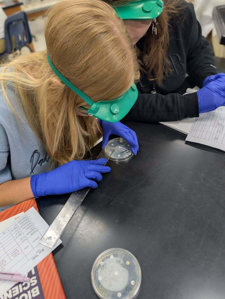 Two individuals, wearing goggles and gloves, examine a petri dish with a substance on a black desk.