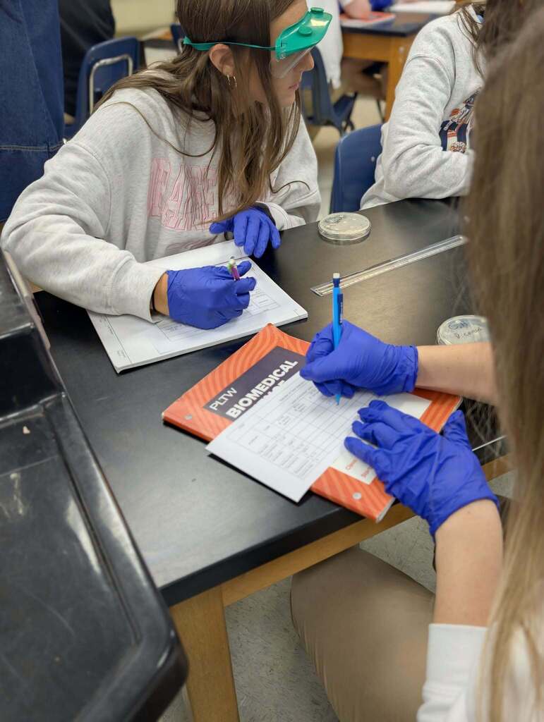 Two students in lab coats and gloves work on a science project. One writes on a paper, and the other holds a pen.