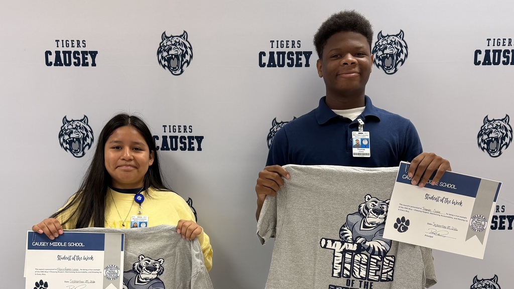 Two individuals hold certificates and gray shirts with tiger emblems in front of a wall.