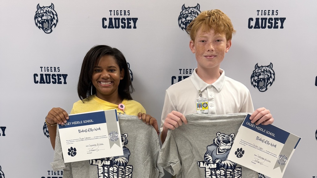 Two kids hold certificates in front of a wall with the word "Tigers" and a tiger logo.