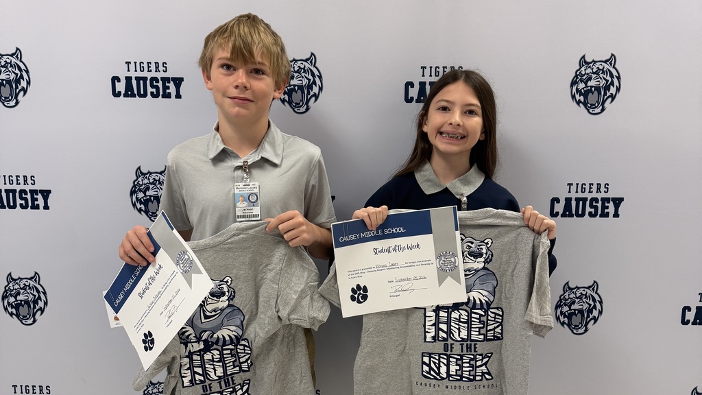 Two kids hold certificates and school shirts in front of a backdrop with a school logo.