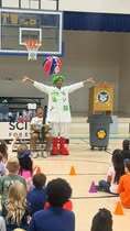 A person in a green costume with a red hat performs for children seated on the floor in a gymnasium.