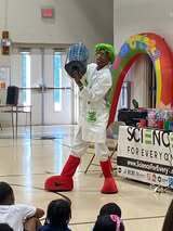 A person in a white suit and red boots performs for a group of children. The room has an inflatable rainbow arch and a sign that reads Science for Everyone.