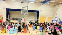 A gymnasium with a basketball hoop. Many children sit on the floor in front of a stage.