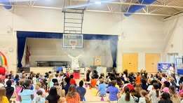 A large indoor auditorium with a stage, a basketball hoop, and children sitting on the floor.