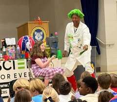 A man in a white lab coat performs a science experiment while a woman sits on a stage. Children sit in front.