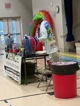 Person in green wig and white coat at a table with a sign reading "Science Performance" and a red trash bin.