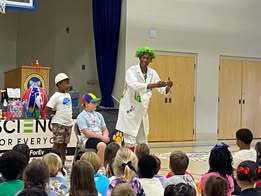 Performers on a stage entertain a group of children in an auditorium. One person wears a lab coat.