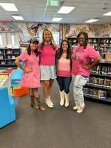 Five women in matching pink tops and white boots stand together in a library. Behind them, bookshelves are filled with books.