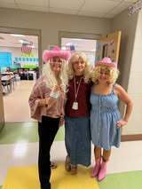 Three adults in wigs and cowboy hats stand in a school hallway. One wears pink, another maroon, and the third blue.