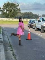 A woman in a pink dress and brown boots stands on the road, near a traffic cone. Cars and a pickup truck are parked on the side.