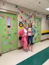 Two individuals stand in a corridor, one in pink and one in blue. Green doors have colorful decorations. White walls, a yellow stripe, and a green floor border.