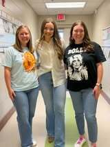 Three women stand together in a hallway. One wears a blue shirt, another a white top, and the last a black t-shirt.