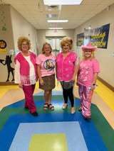 Four adults stand in a hallway, all wearing pink tops. The floor is colorful with a multicolored circle.