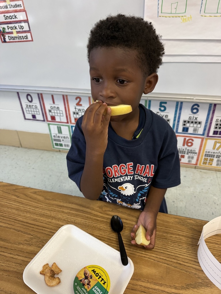 A child eats a banana in a classroom. He holds a plate with crackers. Behind him is a whiteboard with numbers.