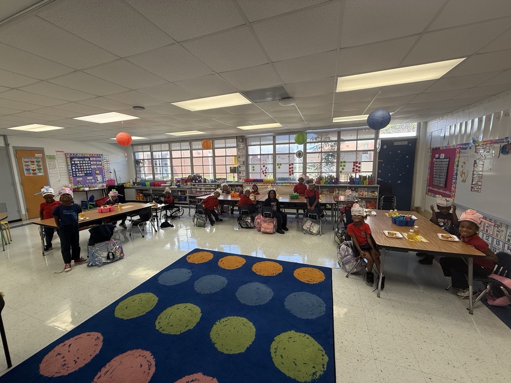 A classroom with students sitting at tables. A multicolored rug with circular patterns is on the floor.