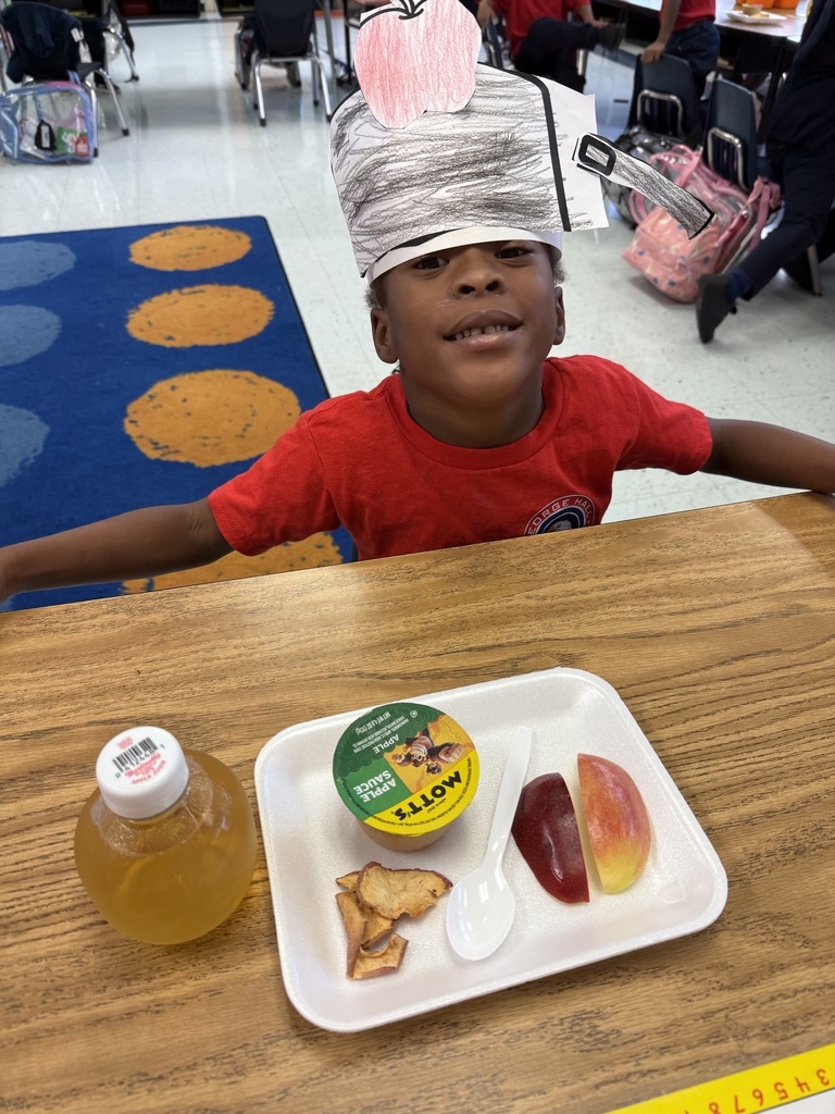 A young child wearing a paper hat sits at a table with a tray of food, including yogurt, apple slices, and crackers.