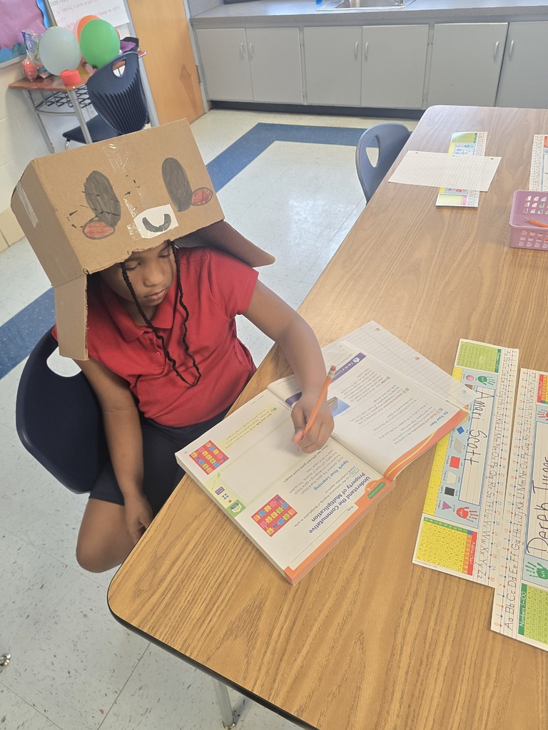 Child wearing a cardboard animal head, seated at a desk writing in a book.