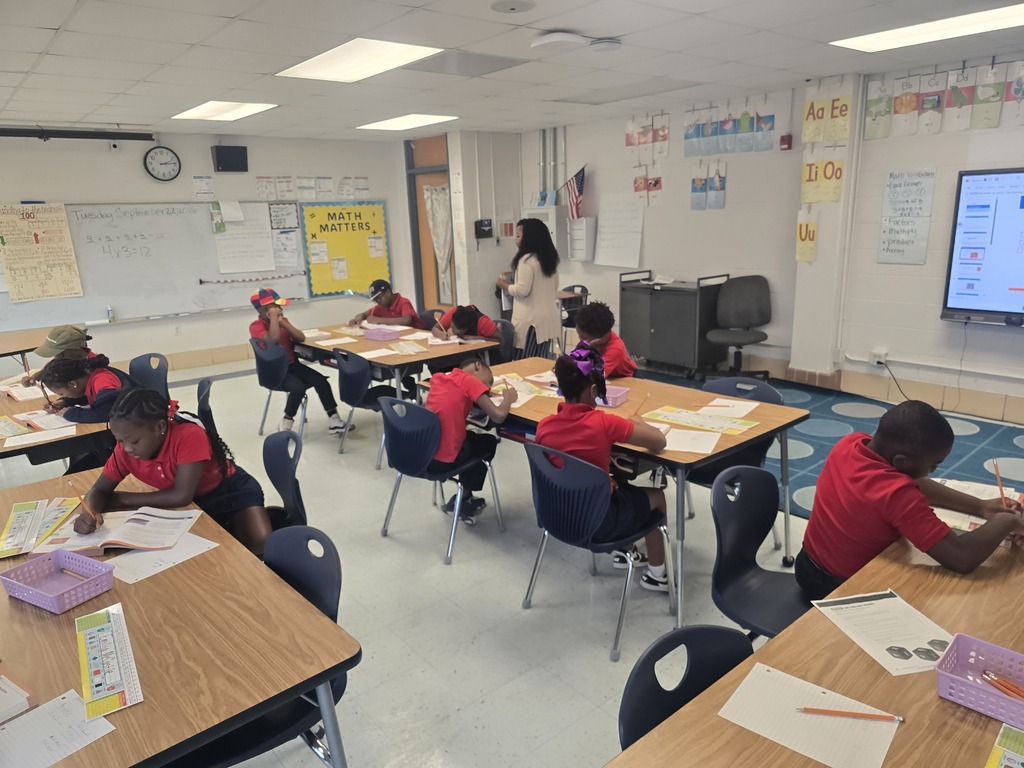Classroom with students in red shirts sitting at desks. A teacher stands near a whiteboard and screen. Bright lighting and bulletin board with posters.
